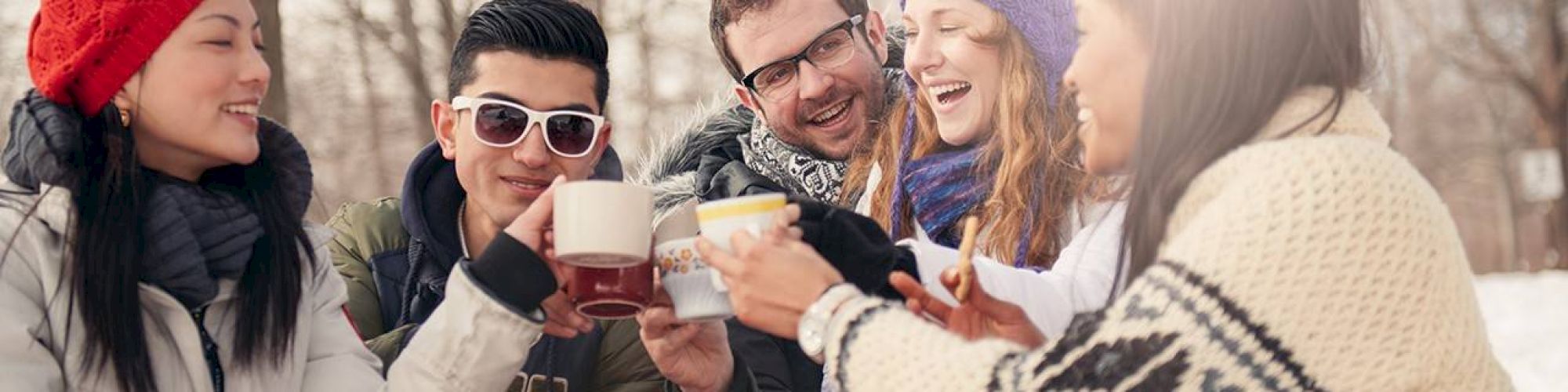 A group of five friends in winter clothes are toasting with mugs outdoors in a snowy setting, smiling and having a good time.