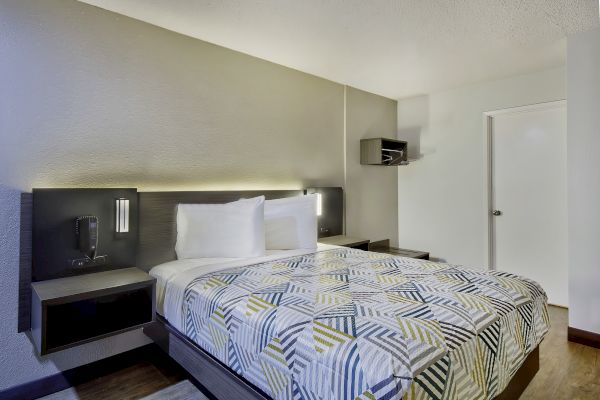 A modern hotel room with a neatly made bed, geometric patterned comforter, nightstands, wall-mounted lights, and a small shelf near the door.