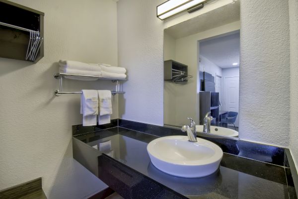 A bathroom vanity with a white sink, dark countertop, towels on a shelf, and a large mirror reflecting a room in the background.