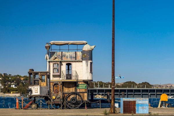 A whimsical, old-fashioned houseboat with an eclectic design is docked at a marina, featuring a mix of traditional and rustic elements.