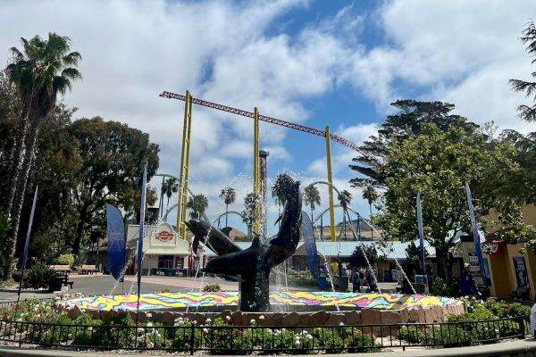 The image shows a public square with a statue in the center, surrounded by colorful decorations, palm trees, and some buildings in the background.