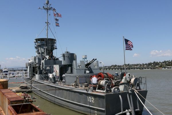 A military ship with the number 102 is docked at a pier, displaying an American flag. The background shows water and a distant shoreline.