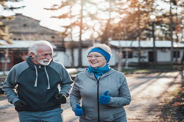 Two elderly people jog together on a tree-lined path with smiles on their faces, enjoying a sunny day outdoors.