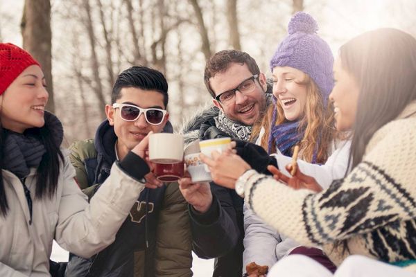 A group of friends wearing winter clothes is outdoors, smiling, and toasting with mugs in a snowy setting.