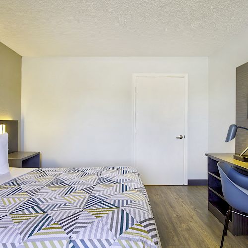 A modern, minimalist hotel room with a neatly made bed, patterned bedspread, desk, chair, lamp, and a wall-mounted TV, ending the sentence.