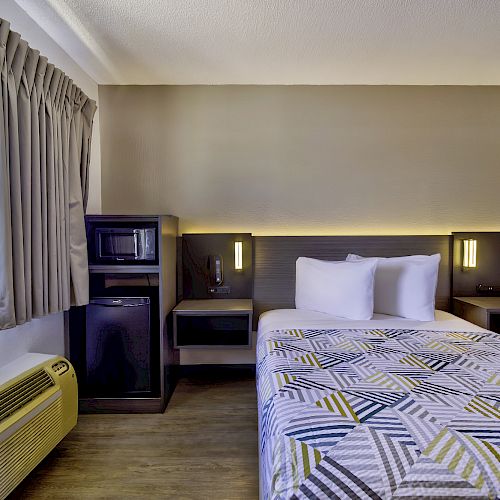 A hotel room features a bed, nightstands, a microwave, fridge, air conditioner, window, and modern decor.