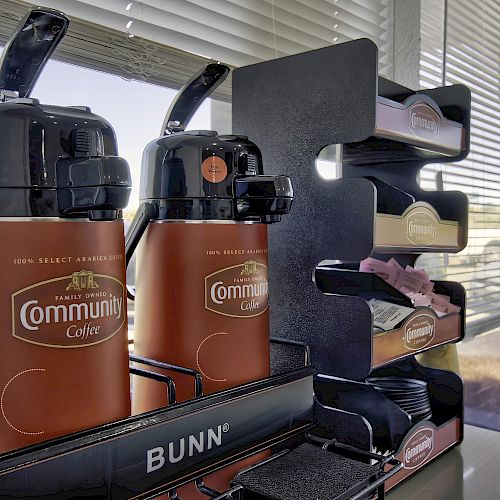 The image shows two Community Coffee dispensers on a Bunn stand and a shelf with coffee accessories like cups and lids near a window. Ending the sentence.