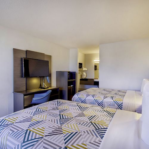 This image shows a modern hotel room with two double beds, a TV on a wall-mounted unit, a desk, and a chair in a clean, minimalist style.