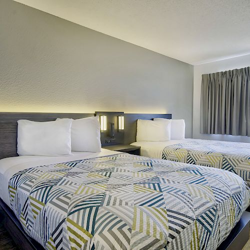 The image shows a hotel room with two neatly made beds, modern bedding, a soft gray wall, a window with curtains, and a wooden floor.