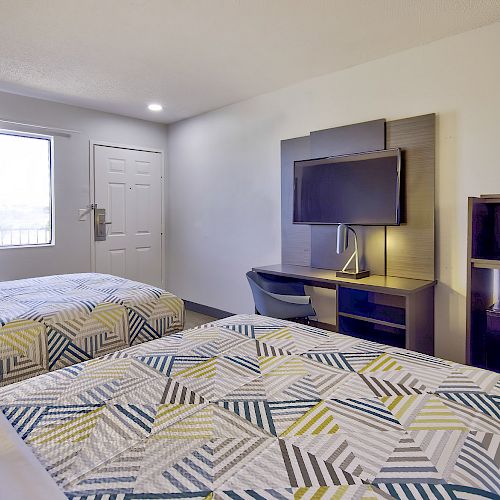 This image shows a hotel room with two beds, a wall-mounted TV, a desk, a microwave, and a refrigerator. The room has a window with curtains.