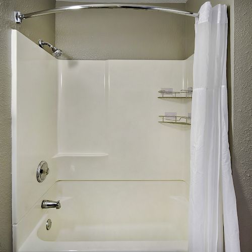 This image shows a clean bathroom shower area with a white bathtub, curtain, and shelves. A towel is hanging on a rod to the left. The walls are beige.