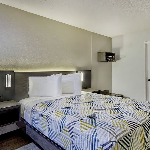 A modern hotel room features a bed with a geometric-patterned bedspread, built-in nightstands, a wall-mounted TV, and a minimalist design.