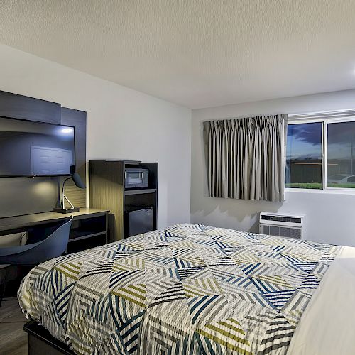 The image shows a cozy hotel room with a large bed, a flat-screen TV mounted on the wall, a desk with a chair, a window with a curtain, and ambient lighting.