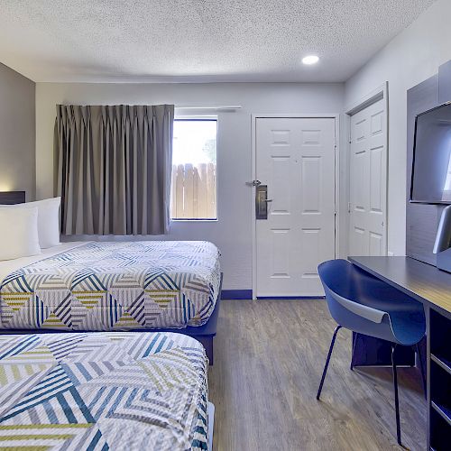 The image shows a clean and modern hotel room with two neatly made beds, a desk with a chair, a television, and a window with curtains.