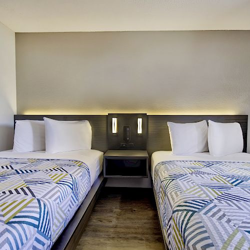 The image shows a hotel room with two single beds, each with geometric-patterned bedspreads, pillows, and a central nightstand with two lights.