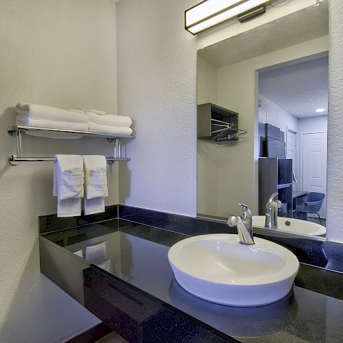 A modern bathroom with a white sink on a black countertop, a wall-mounted shelf with towels, and a large mirror reflecting more of the room's interior.