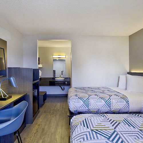 This image shows a hotel room with two beds, a desk, chair, TV, and a bathroom sink area facing the beds, all in modern decor.