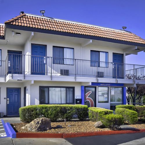 The image shows a two-story motel with a tiled roof, blue doors, and balconies on the upper level, surrounded by bushes and a 