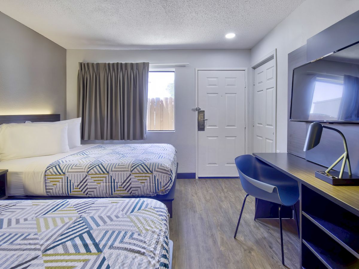 The image shows a well-lit hotel room with two beds, a desk, a chair, a flat-screen TV, and a window with curtains.