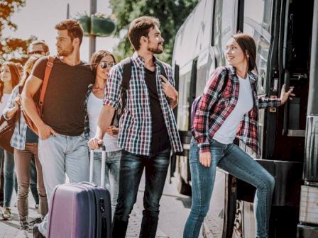 A group of young people are standing in line to board a bus, with luggage and backpacks, enjoying the outdoor scene and chatting together.