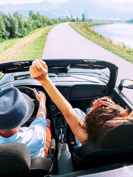 Two people are driving in a convertible on a scenic road by a lake, with the passenger raising their arms joyfully.